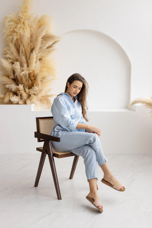 Woman sitting in a wooden chair, dressed in dusty blue linen pants and a light blue knotted blouse, exuding relaxed, casual elegance in a bright, airy setting.