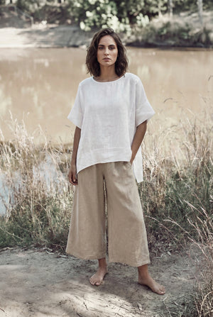 Model standing barefoot by a river wearing a breezy white linen tunic top with a relaxed fit.