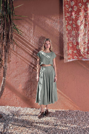 Model wearing the NAOMI dusty sage linen two-piece set, featuring a boxy crop top and high-waisted midi skirt, standing outdoors against a warm terracotta wall.