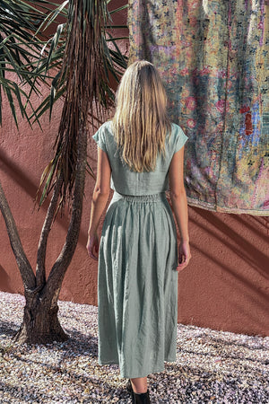 Back view of the model wearing the NAOMI dusty sage linen two-piece set, highlighting the relaxed crop top and the elastic back waistband of the high-waisted midi skirt.