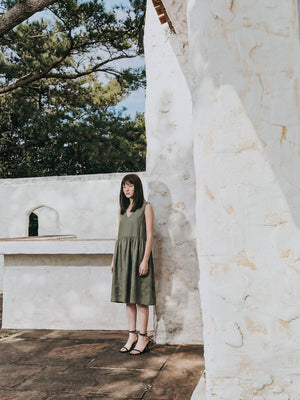 Full-length view of a model styled in the EMILIJA eucalyptus green linen sleeveless midi dress with black strap heels, standing outdoors beside a white stone structure with green trees in the background.