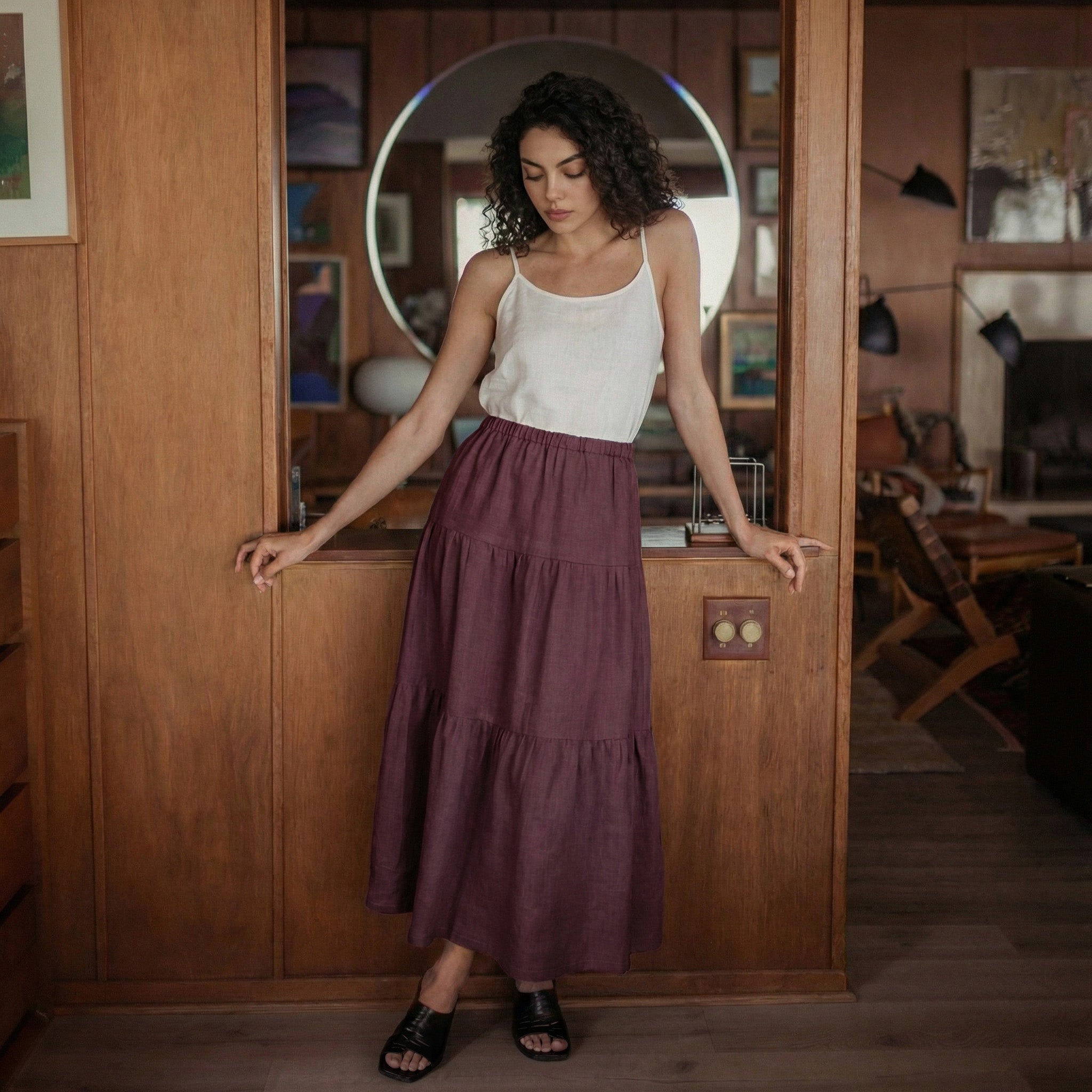 Woman standing in a wooden doorway wearing a grape wine tiered linen skirt and white camisole top.