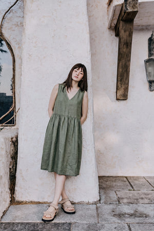 oman wearing the EMILIJA eucalyptus green linen sleeveless midi dress with a V-neckline and a relaxed smock silhouette, leaning against a textured light-colored stucco wall.