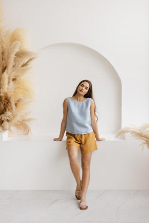Model lounging in mustard linen shorts against a white arch backdrop with natural decor.