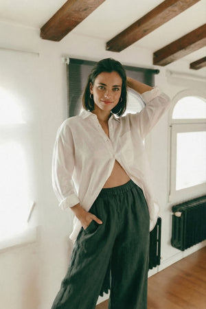 Woman posing casually in MINA smoke pine wide leg linen pants, highlighting the comfortable elastic waistband and deep pockets, paired with an unbuttoned white shirt.