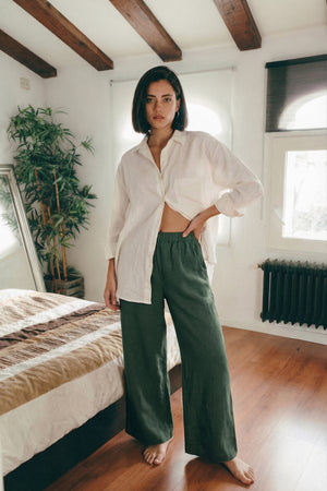 Woman wearing MINA wide leg linen pants in smoke pine green, styled with a relaxed white linen shirt in a bedroom setting.