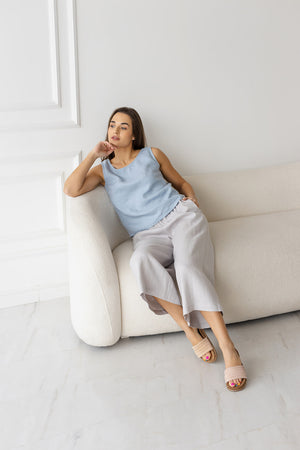 Woman lounging on a cream sofa wearing a light blue sleeveless linen top paired with flowy grey wide-leg pants in a minimalist interior.