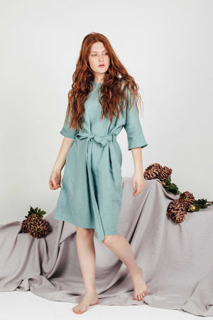 Front view of a breathable sea-green linen boho dress, featuring a knee-length hem, side pockets, and ¾ sleeves, styled barefoot against a draped backdrop.