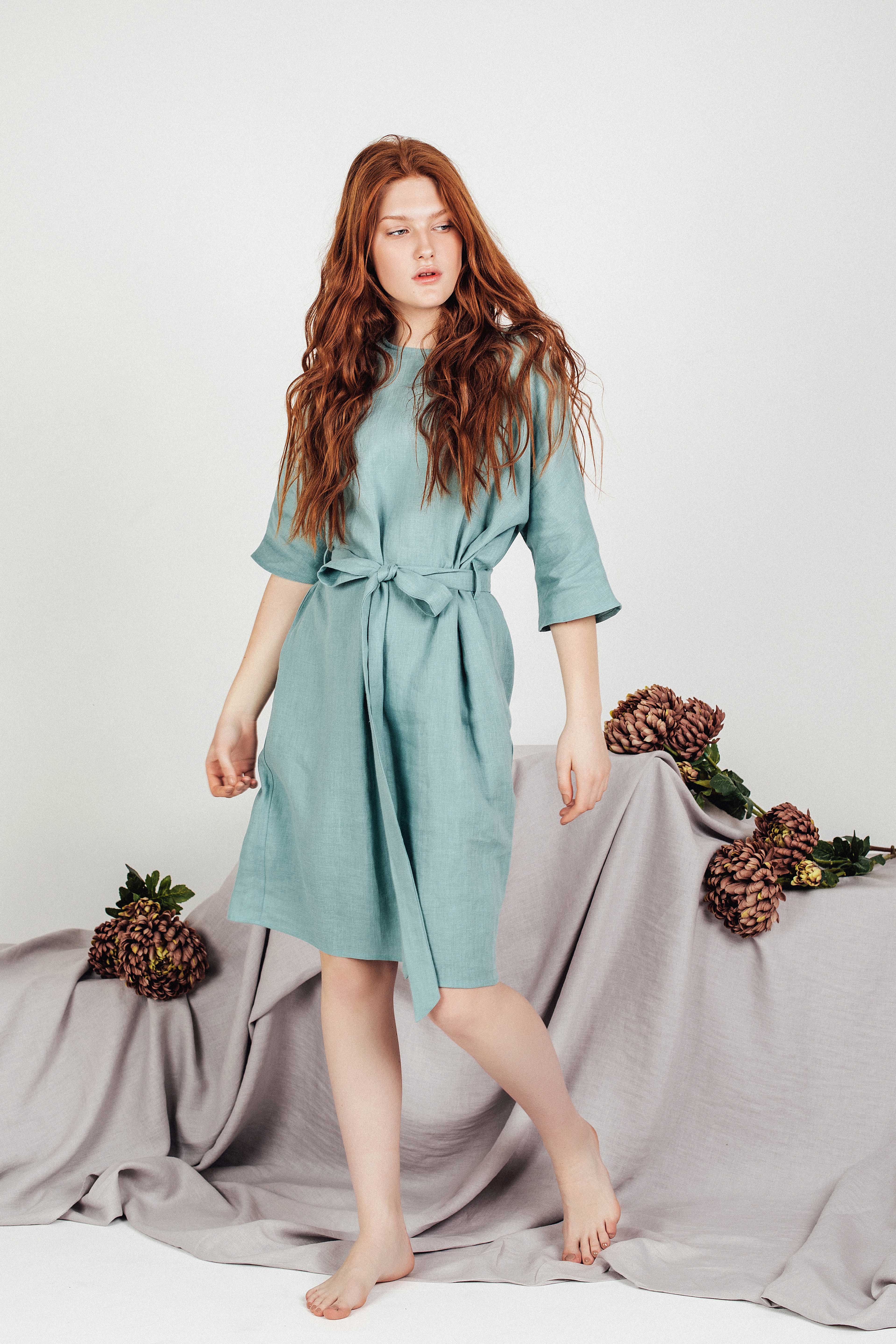 Front view of a breathable sea-green linen boho dress, featuring a knee-length hem, side pockets, and ¾ sleeves, styled barefoot against a draped backdrop.