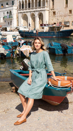 Model wearing a breathable sea-green knee-length linen dress, sitting on a small wooden boat with a sunny Mediterranean harbor in the background.