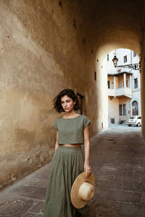 Model wearing the Sonfre NAOMI eucalyptus linen two-piece set, featuring a boxy crop top and a high-waisted midi skirt, holding a straw hat while walking through a stone alleyway.