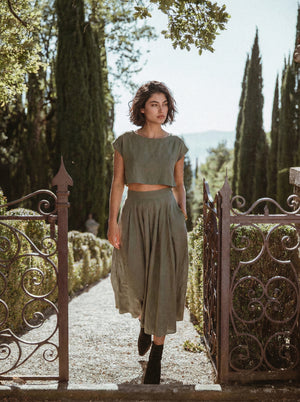 Model wearing the Sonfre NAOMI eucalyptus green linen two-piece set, featuring a boxy crop top and a high-waisted midi skirt, walking through an ornate iron gate in a garden setting.