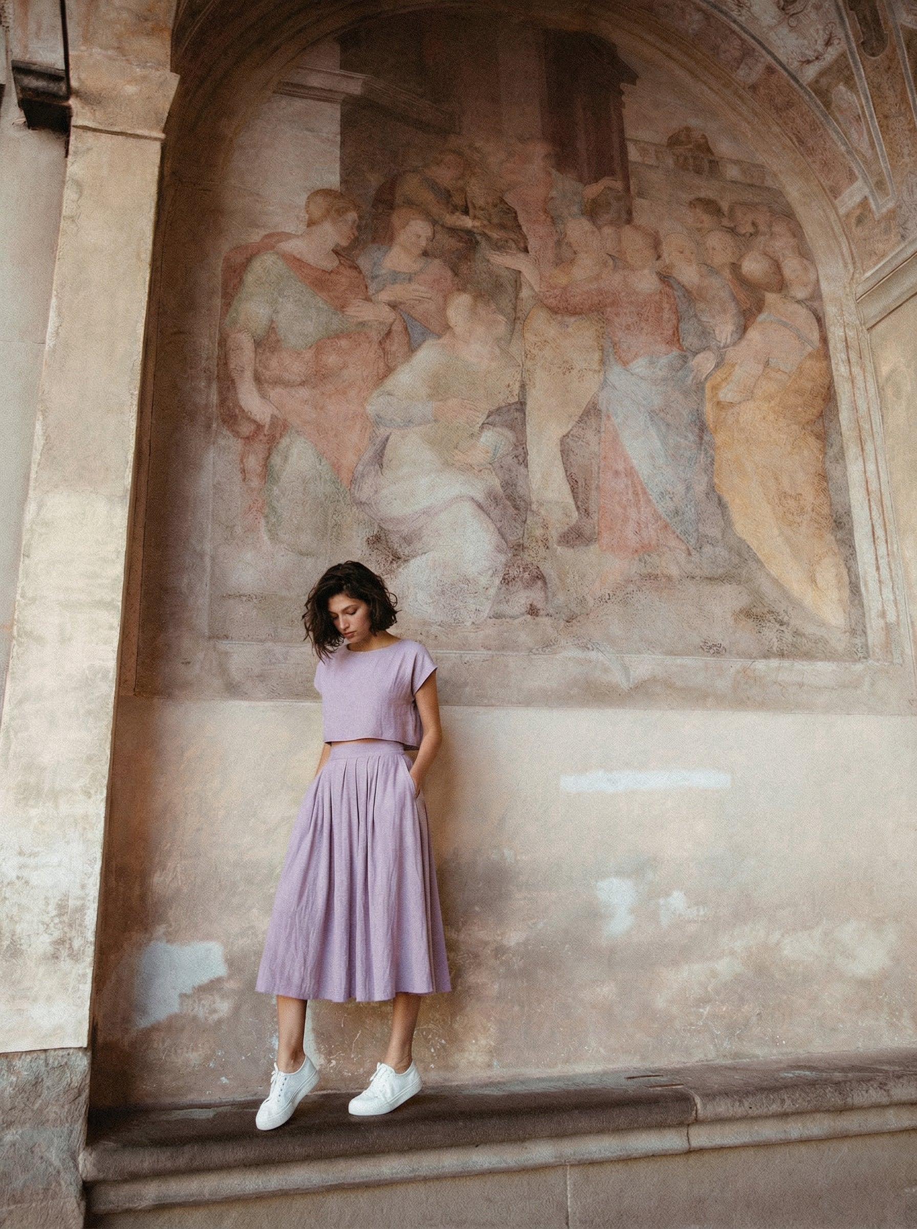 Model wearing the Sonfre NAOMI lavender linen short-sleeve crop top and matching high-waisted midi skirt with pockets, standing in front of a classical stone mural.