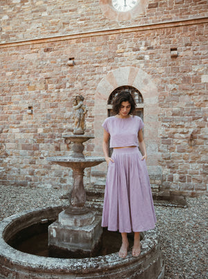 Model wearing the Sonfre NAOMI lilac linen two-piece set, featuring a cropped top and a high-waisted midi skirt, standing next to an old stone fountain against a brick wall.