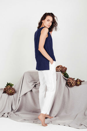 Woman standing barefoot in a sleeveless deep navy linen top and white linen pants, facing sideways with one hand in her pocket, in front of neutral draped fabric and dried florals.