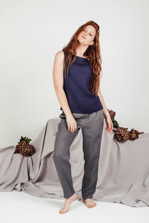 Red-haired model wearing a navy linen top with grey pants, leaning slightly with one leg forward, barefoot in a calm, minimalist scene featuring neutral drapery and florals.