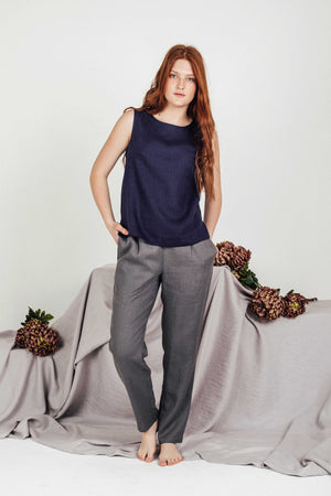 Red-haired woman in a navy blue linen sleeveless top and high-waisted grey pants, barefoot with a soft expression, styled against a neutral draped fabric backdrop with dried flowers.