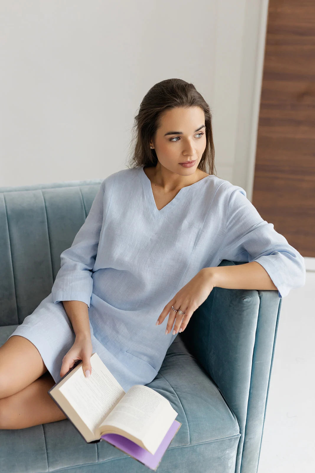 Woman wearing a light blue handmade linen dress, sitting on a velvet couch reading a book – Sonfre sustainable fashion