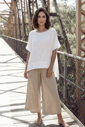 Woman standing barefoot on a sunlit bridge wearing an oversized white linen short-sleeve tunic top and wide-leg beige linen pants.