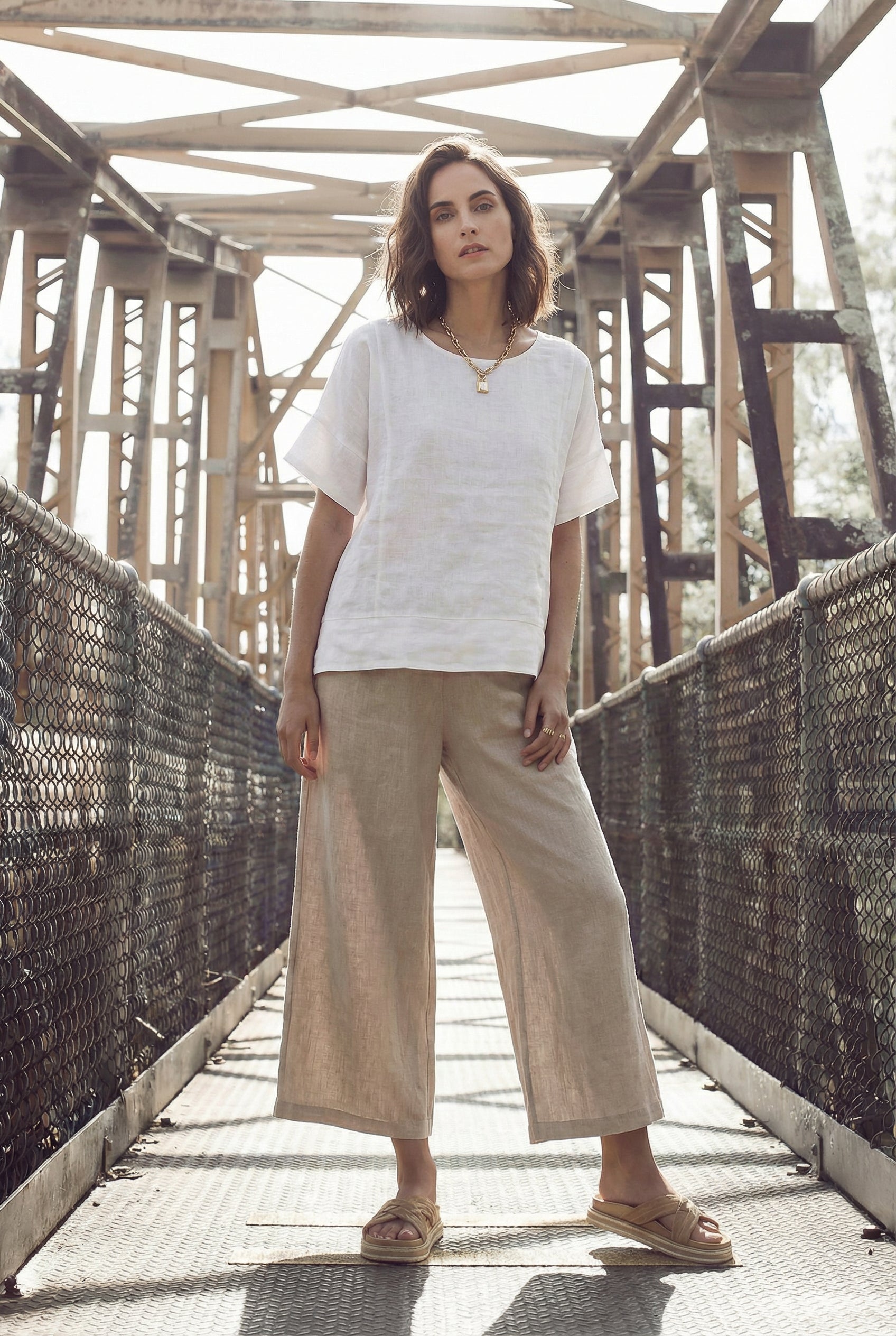 Model wearing a loose white linen tunic top with a high-low hem, standing on a bridge.