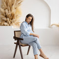 Woman sitting in a wooden chair, dressed in dusty blue linen pants and a light blue knotted blouse, exuding relaxed, casual elegance in a bright, airy setting.