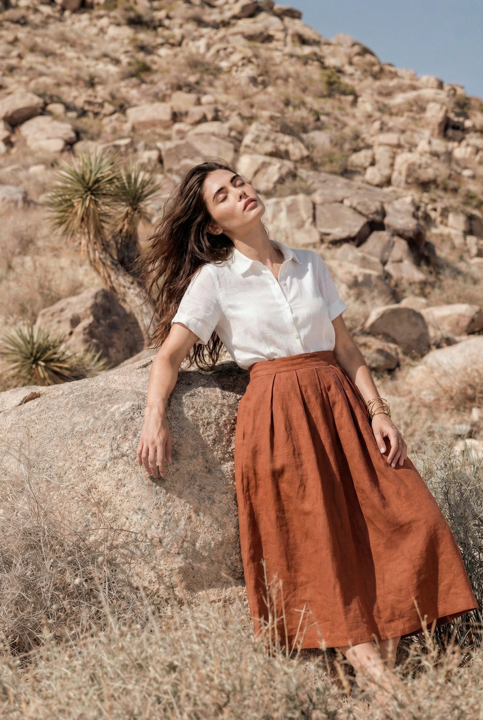 Woman leaning against a large boulder in a desert landscape, modeling the Aivy Classic Linen Midi Skirt in Rust paired with a white short-sleeve blouse.
