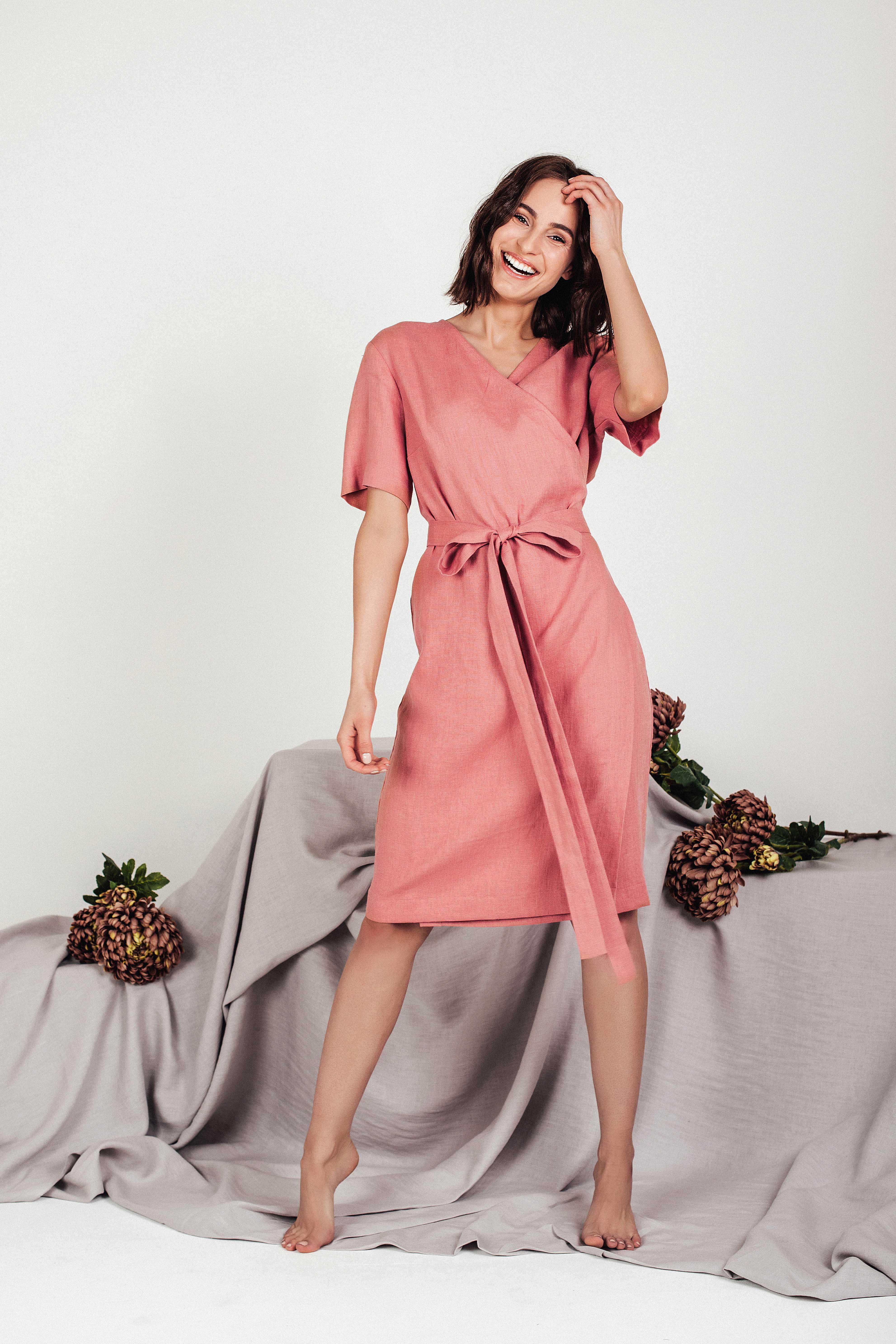 Model in a blush pink linen wrap dress with a V-neck, short sleeves, and tie waist, perfect for casual summer days.