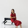 Model wearing a burgundy red linen boho dress with a relaxed, flowy fit, sitting on a chair in a minimalist setting.