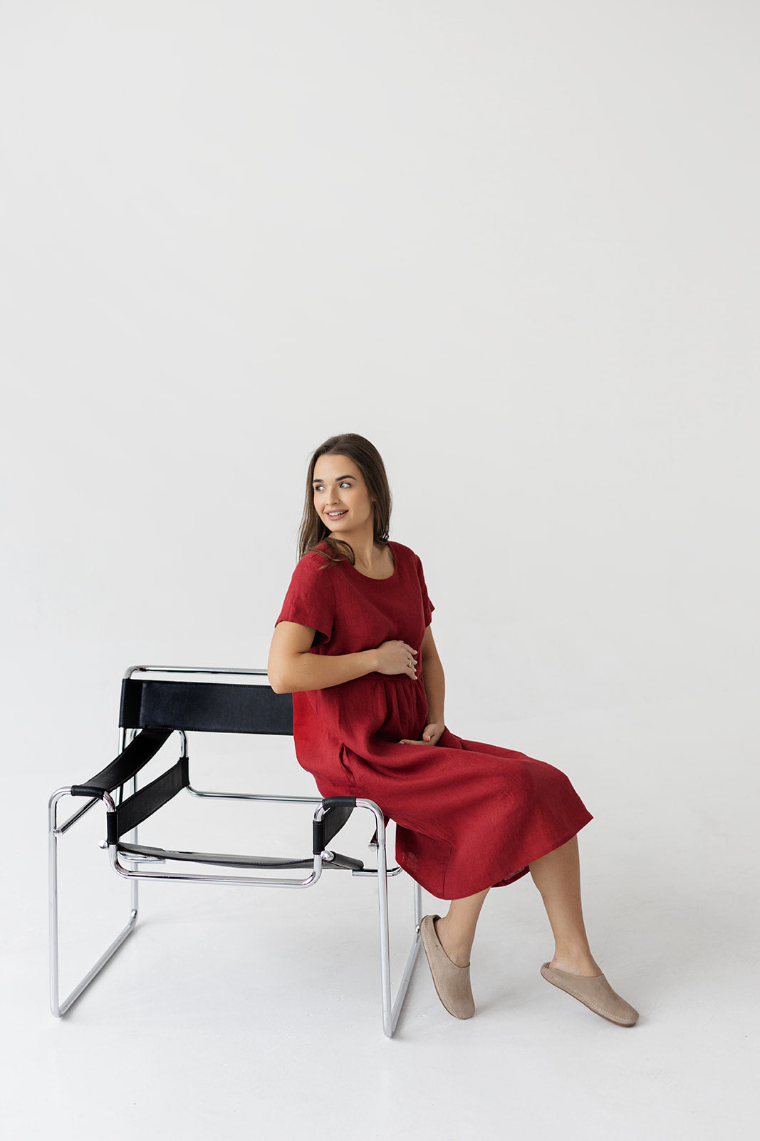 Model wearing a burgundy red linen boho dress with a relaxed, flowy fit, sitting on a chair in a minimalist setting.