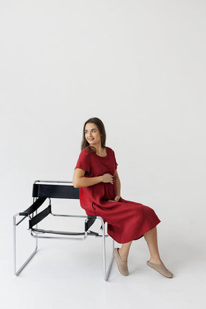 Model wearing a burgundy red linen boho dress with a relaxed, flowy fit, sitting on a chair in a minimalist setting.