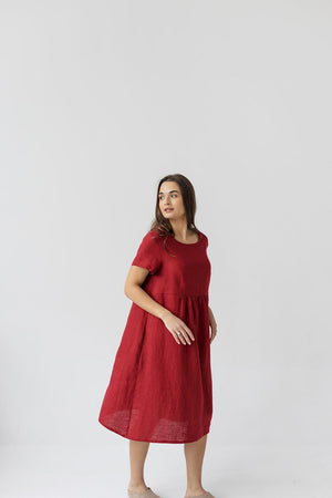 Full-length view of a model standing, wearing a burgundy red linen maternity dress with a wide scoop neckline. The dress drapes loosely, demonstrating its comfortable, relaxed fit.