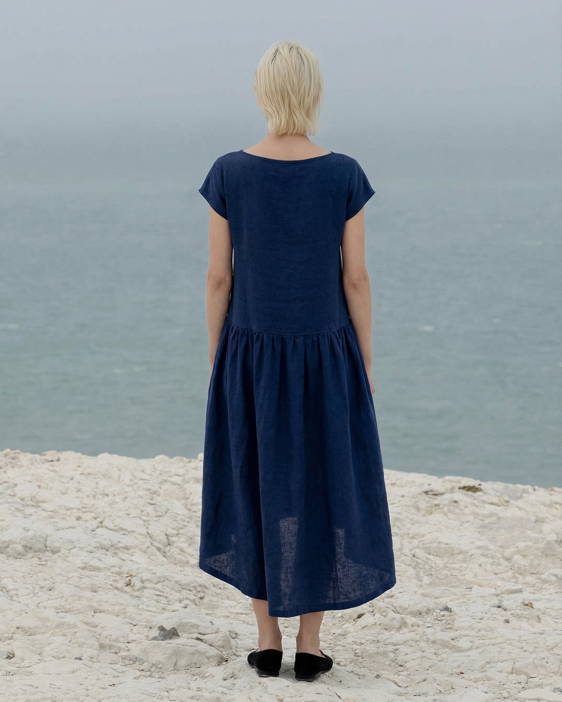Back view of a woman wearing the 2026 Daiva midnight blue linen smock dress, highlighting the gathered waistline and relaxed midi fit as she looks out over the ocean.