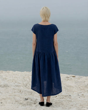 Back view of a woman wearing the 2026 Daiva midnight blue linen smock dress, highlighting the gathered waistline and relaxed midi fit as she looks out over the ocean.
