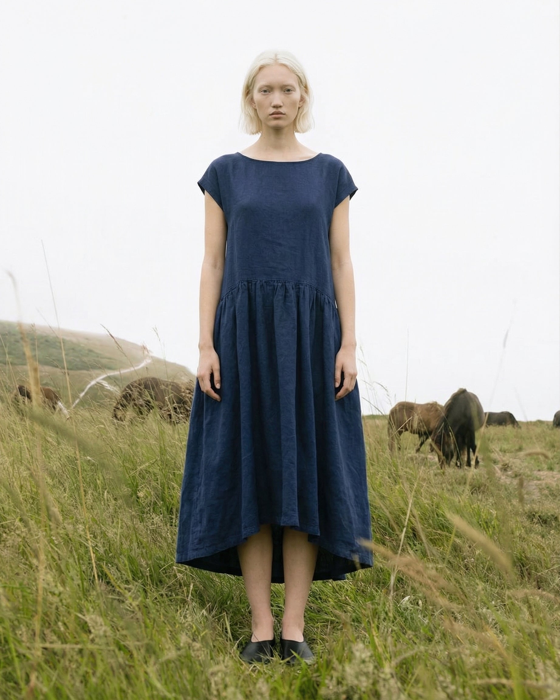 Full front view of a woman wearing the 2026 Daiva midnight blue linen smock dress, standing in a grassy coastal field with wild horses grazing in the background.