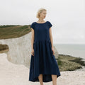 Full body view of a woman wearing the Daiva midnight blue linen smock midi dress with a gathered waist, standing on a white coastal cliff.