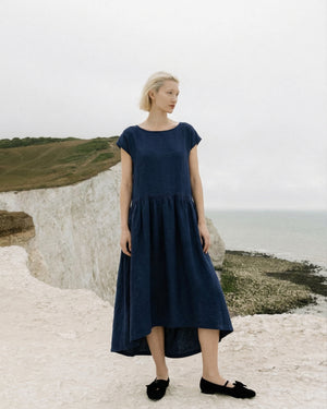 Full body view of a woman wearing the Daiva midnight blue linen smock midi dress with a gathered waist, standing on a white coastal cliff.