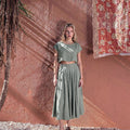 Model wearing the NAOMI dusty sage linen two-piece set, featuring a boxy crop top and high-waisted midi skirt, standing outdoors against a warm terracotta wall.