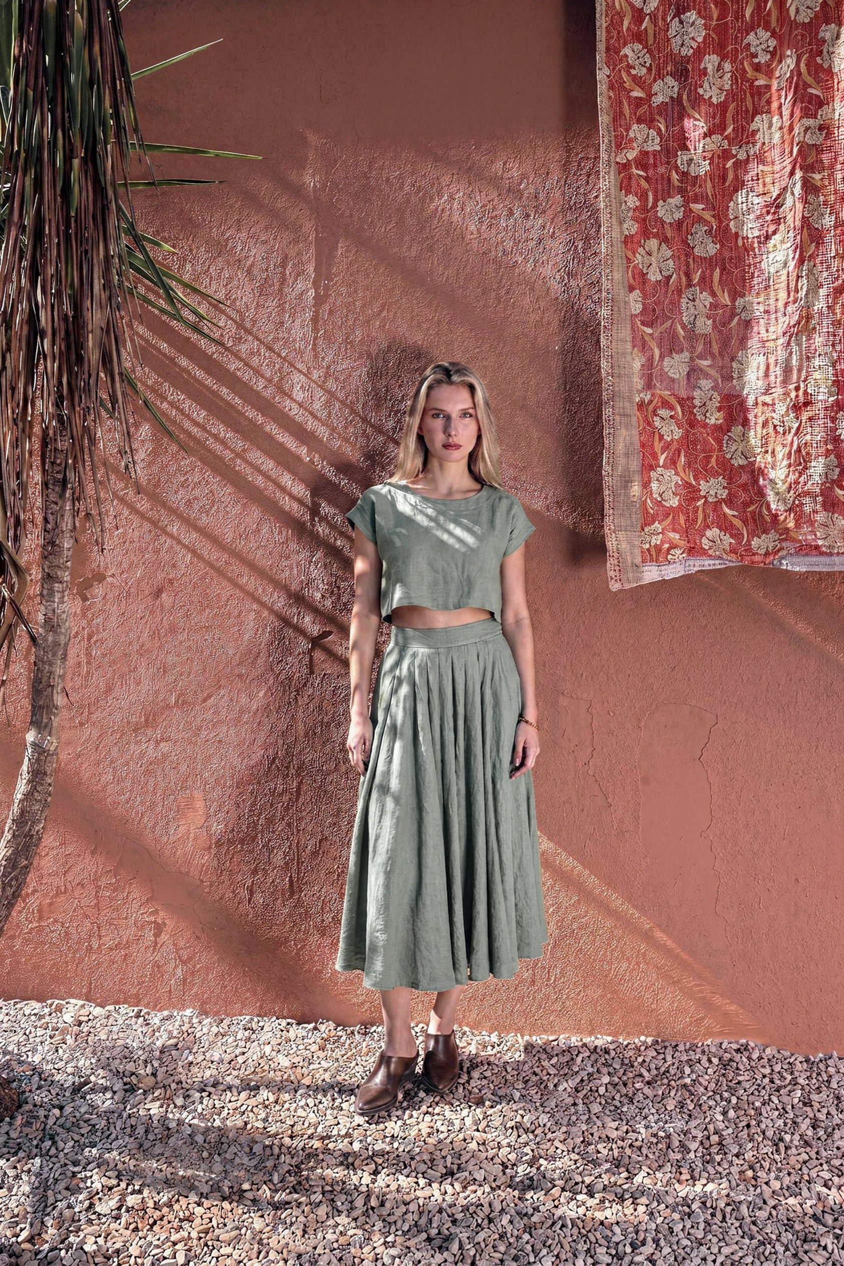 Model wearing the NAOMI dusty sage linen two-piece set, featuring a boxy crop top and high-waisted midi skirt, standing outdoors against a warm terracotta wall.