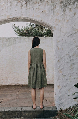 Back view of the EMILIJA eucalyptus green linen sleeveless midi dress, highlighting the clean high-back neckline and the gathered waist design as the model stands under a stone archway.