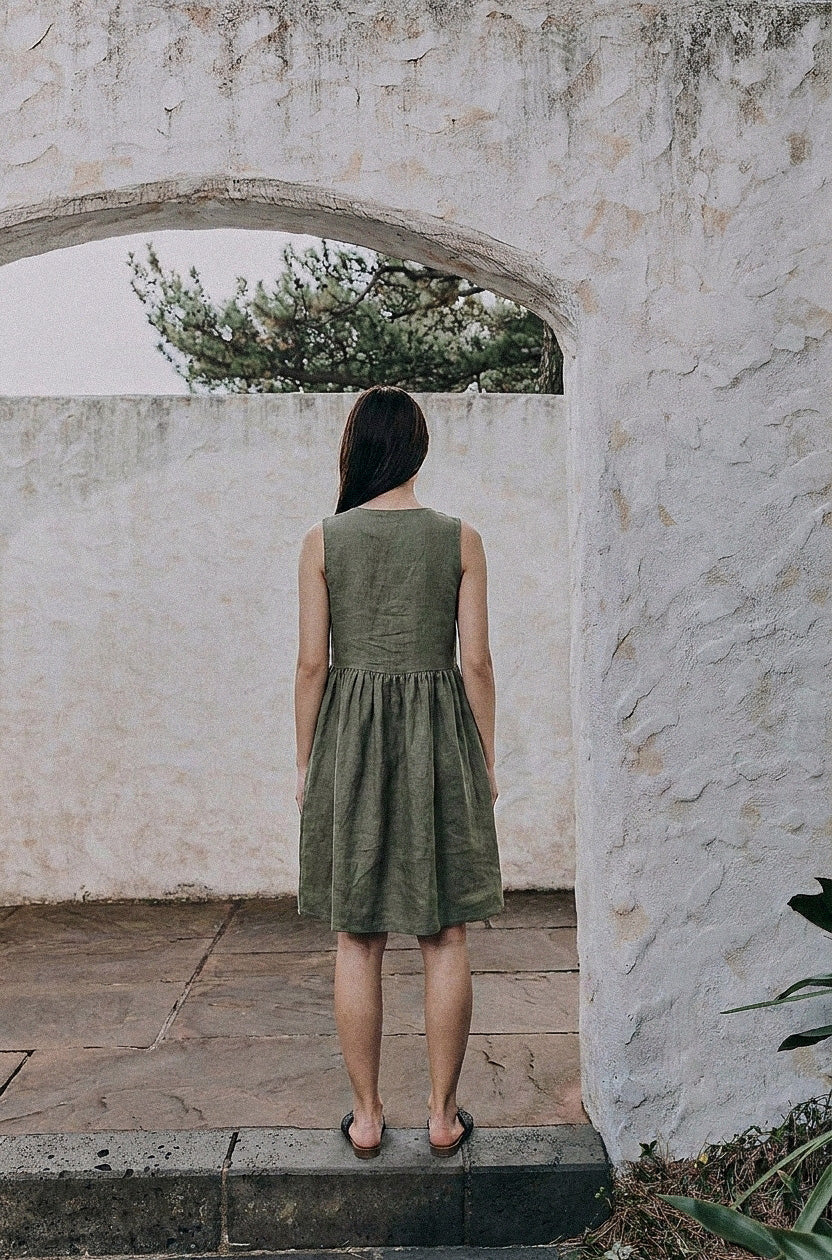 Back view of the EMILIJA eucalyptus green linen sleeveless midi dress, highlighting the clean high-back neckline and the gathered waist design as the model stands under a stone archway.