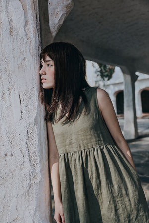 Close-up side view of a woman wearing the EMILIJA eucalyptus green linen sleeveless dress, showing the texture of the linen fabric and the V-neckline detail as she leans against a textured plaster archway.