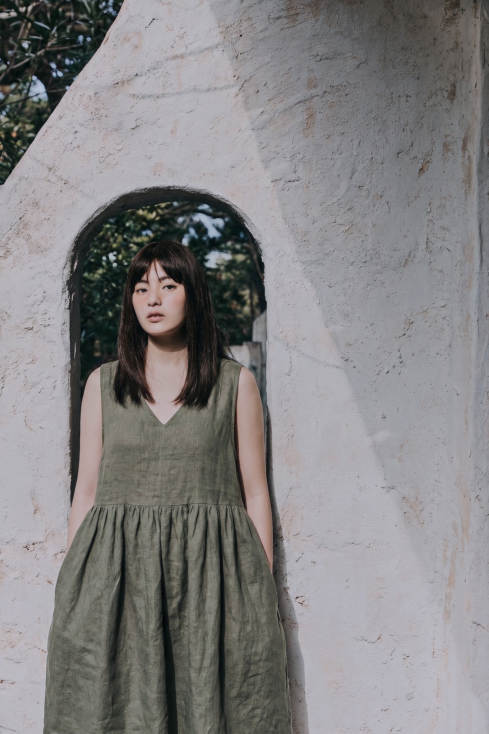 A front-facing portrait of a woman wearing the EMILIJA eucalyptus green linen sleeveless midi dress, showcasing the V-neckline and gathered waist, framed inside a rustic stone stucco archway.