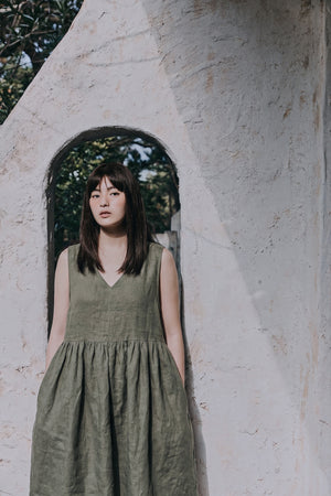 A front-facing portrait of a woman wearing the EMILIJA eucalyptus green linen sleeveless midi dress, showcasing the V-neckline and gathered waist, framed inside a rustic stone stucco archway.