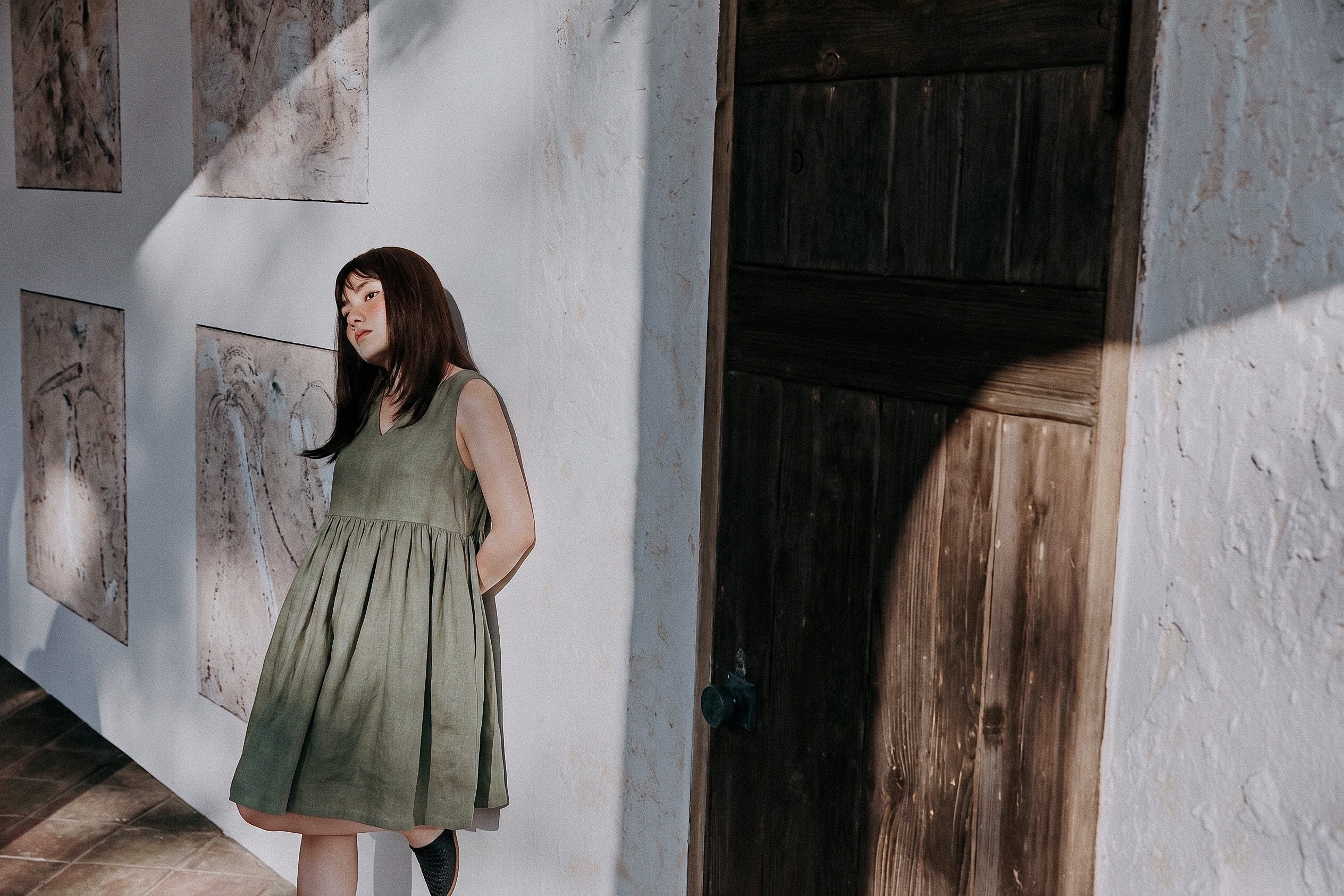 A lifestyle shot of a woman in the EMILIJA eucalyptus green linen sleeveless dress leaning against a light stucco wall next to a rustic wooden door under warm afternoon sunlight.