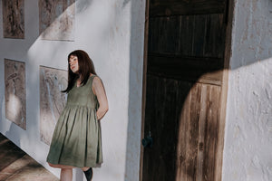 A lifestyle shot of a woman in the EMILIJA eucalyptus green linen sleeveless dress leaning against a light stucco wall next to a rustic wooden door under warm afternoon sunlight.