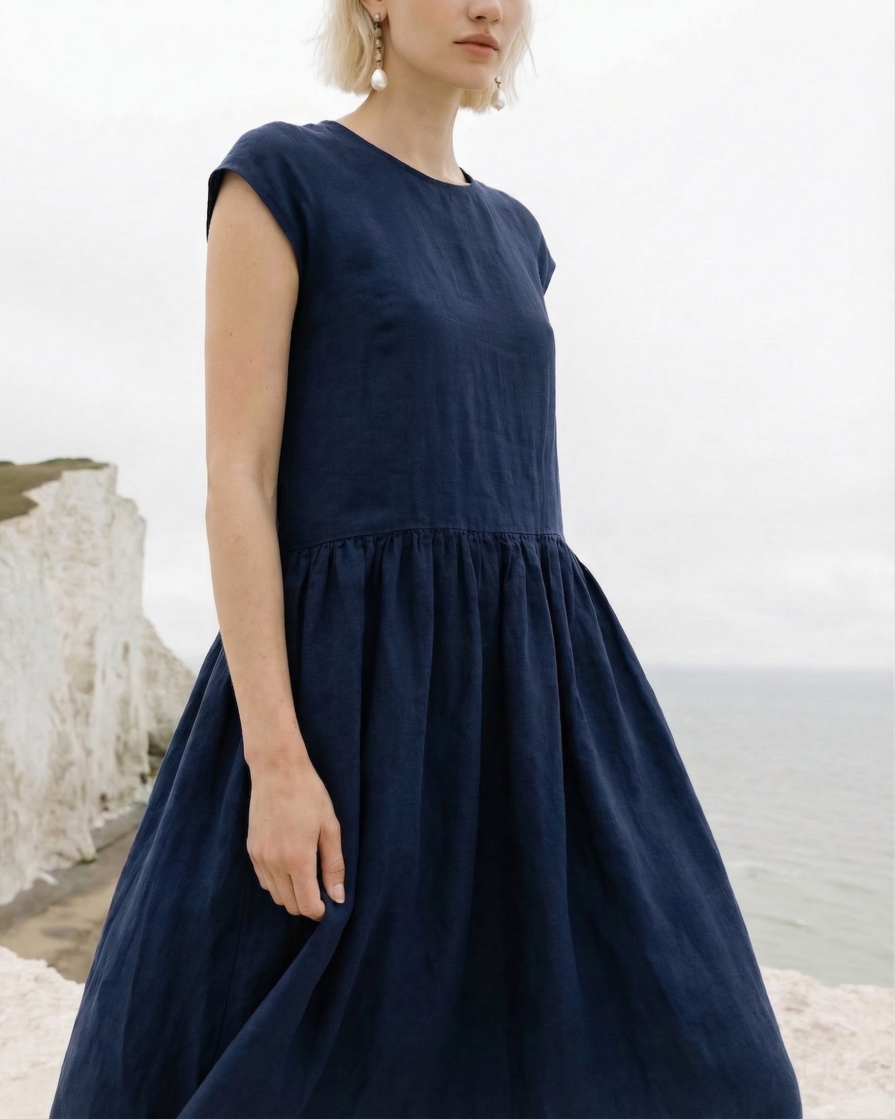 Close-up of the gathered mid-waist seam and flowing skirt on the Daiva midnight blue linen smock dress, worn by a model standing near white coastal cliffs.
