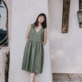 oman wearing the EMILIJA eucalyptus green linen sleeveless midi dress with a V-neckline and a relaxed smock silhouette, leaning against a textured light-colored stucco wall.