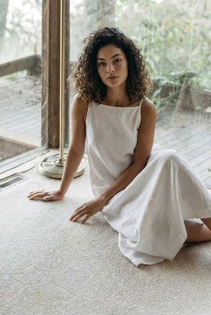 Woman sitting on floor wearing breathable white linen summer dress, showing A-line skirt flow.