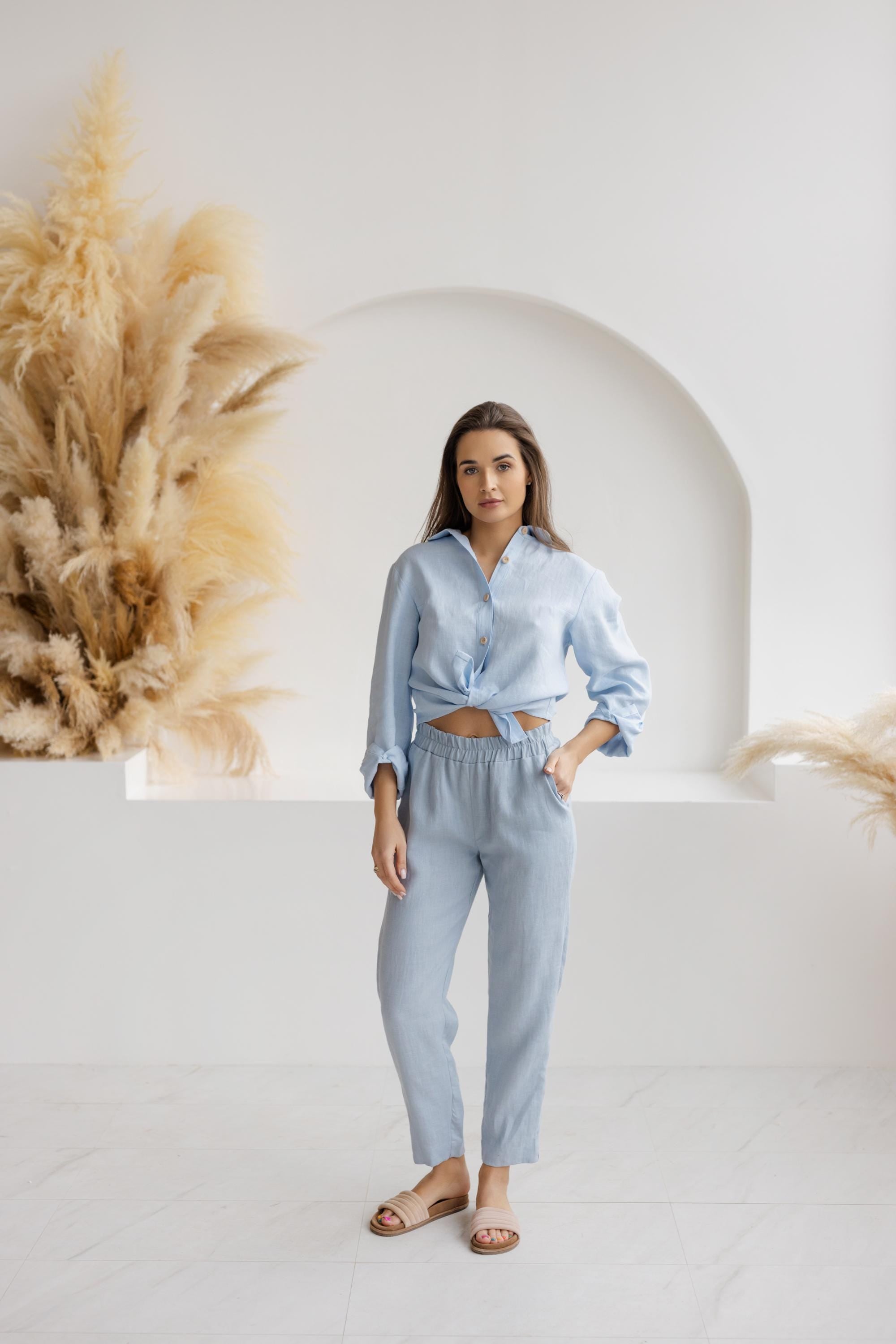 Woman wearing relaxed-fit dusty blue linen pants with an elastic waistband and a light blue knotted blouse, standing in a minimalist white space with pampas grass decor