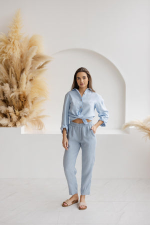 Woman wearing relaxed-fit dusty blue linen pants with an elastic waistband and a light blue knotted blouse, standing in a minimalist white space with pampas grass decor